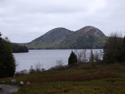 [Two round-top mountains right beside each other looking like the back of a two humped camel sit beside a lake. The sky is completely overcast which helps outline the lumps of the mountains.]